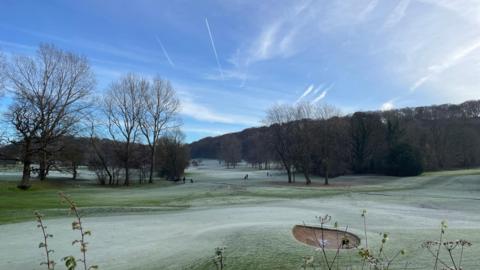 A frosty golf course with an expanse of trees either side under a blue sky with wispy clouds