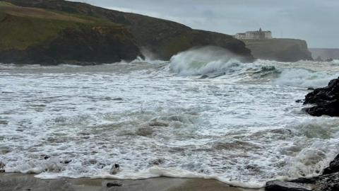 Waves crash in at the coast near Gunwalloe, Cornwall. The sea is white with the foam with sand in the foreground and two headlands in the background - on the further away one are buildings - white or cream with dark roofs