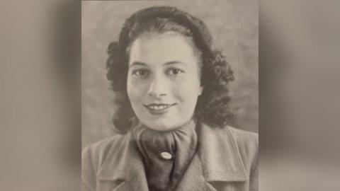 A black-and-white photograph of a young woman with curly shoulder-length brown hair and a hairband keeping her hair back. She is wearing a scarf and a coat and is smiling at the camera.