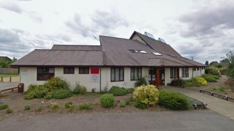 Red Lodge Millennium Centre near Newmarket in Suffolk. It shows a large white building with a grey roof. There are numerous windows and a large entrance. Shrubs and bushes surround the outside of the building.