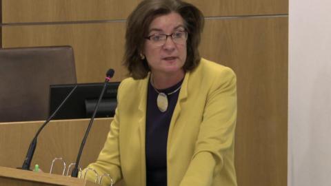 Eluned Morgan stood in the Senedd debating chamber wearing a canary yellow jacket and black top, with glasses, speaking to her left with microphones in front of her.