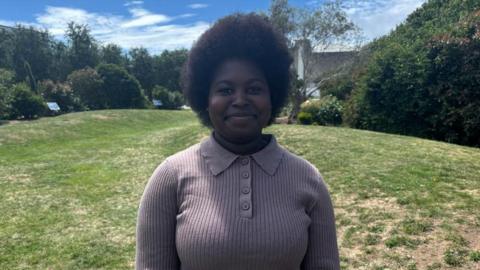 Zahara Noho is smiling as she stands in a park and with grass and shrubs behind. It is a sunny day and there is a wisp of cloud in the sky.