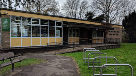 The outside of Chertsey Library, showing the sign on the door, with a park bench in front of the building, along with bicycle racks.