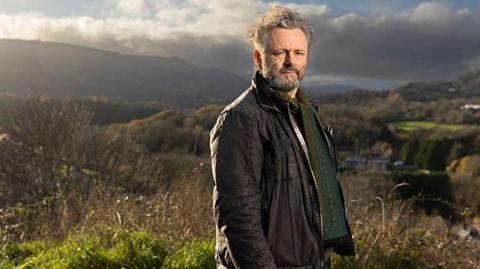 Standing on high ground above a valley, Michael Sheen looks towards the camera with rolling hills and patchwork fields stretching into the distance. He is wearing a brown jacket with a green patterned jumper. Sheen has grey hair and facial hair. Strong afternoon sunlight and heavy clouds create dramatic contrasts across the rural landscape.