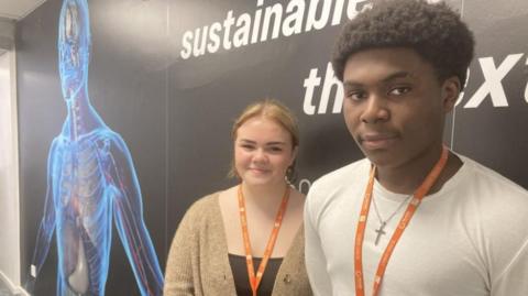 A woman with blond hair, a light brown cardigan and an orange lanyard alongside a man with black hair a white tshirt and an orange lanyard, stood in front of a black wall with a picture of a person's internal organs underneath a blue outline