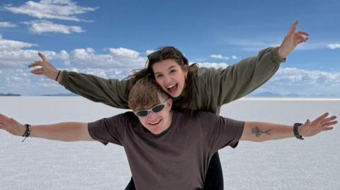 Sioned is pictured on Fin's back. They are both waving their arms in the air. They are pictured at the Uyuni Salt Flats in Bolivia. Fin is wearing sunglasses and they are both smiling.