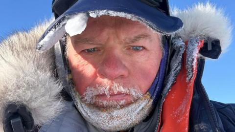 Selfie of Ian Hughes in Antartica. He has small icicles hanging from his moustache and his beard has frozen droplets. He wears a blue cap with ice frozen on the rim and a fur-lined hooded coat over layers.