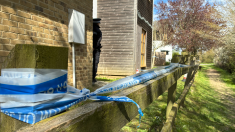 Blue and white police tape runs along the top of a fence beside a house. A police officer with his back to the camera stands next to a house.