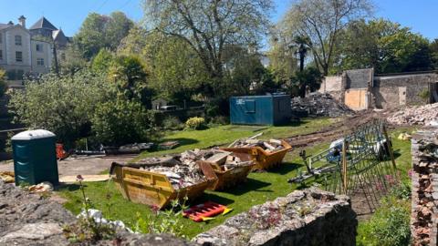 The Singleton Gardens site with a portable toilet container in the left, three skips filled with building materials and a metal shipping container in the background.