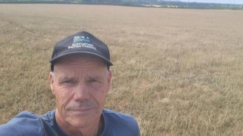 Duncan Palmer, a middle-aged man wearing a baseball cap is stood in a field of yellow grass.
