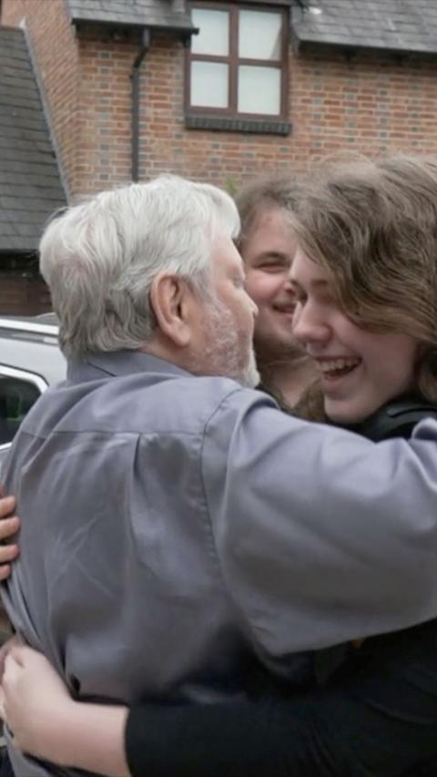 A man wearing a grey shift with grey hair hugging two young men with long hair. They are all smiling