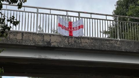 A St George's flag with the word 'England' across it flies on a concrete bridge above a road. 