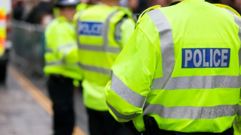 Three police officers facing away from the camera - including two who are out of focus and standing in the distance. The image is taken from behind and the word POLICE can be seen on a hi-vis jacket being worn by one of the officers.