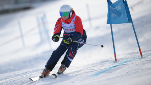 Annabelle Lamb ski ing on a snowy slope, with a blue flag to the right of the image. She is wearing navy trousers with GREAT BRITAIN on the thigh, an orange top, white helmet and blue/green goggles.