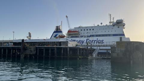 An image of a blue and white Brittany Ferries boat at the Guernsey Port, which has stairs and pillars leading up from the ocean and 'Welcome to Guernsey' written on the side.