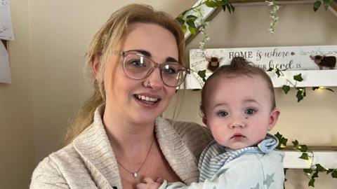 Kiely Luscombe pictured with blonde hair tied back in a pony tail. She is wearing glasses and a beige cardigan with a black top underneath. She is holding Arnold who is wearing a blue baby grow with stars on.