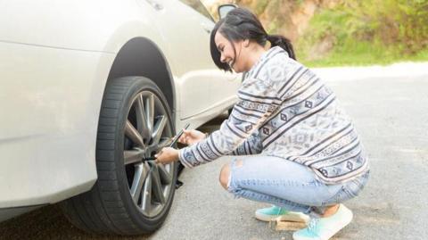 A young woman stranded on the side of the road deals with a flat tyre. She is crouching by a wheel and holding a gadget. She is wearing jeans and a jumper and trainers, with her hair in a ponytail.