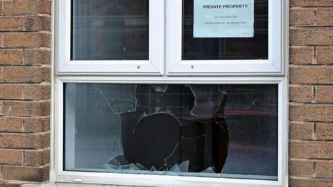 A smashed window is in-between two sides of a brick wall. The window is white. It has a sign on the top right segment saying private property. 