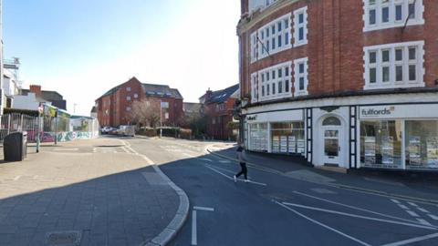 A man walks across the entrance to Gibbon Lane with a shop front on the right side and flats further up the street. 