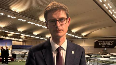 A man stands in a leisure centre where tables are in the background for the vote count. He has a navy suit, burgundy tie, brown short hair and black square glasses. He has a neutral expression but looks sad.