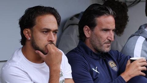 A young footballer with slick black hair wearing a white t-shirt is sat next to a man in his late 30s who is wearing a dark blue jacket with the Notts County logo on it, holding a hot drink.