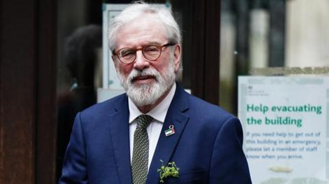 Adams is wearing a navy suit and has a bunch of green shamrocks in the top pocket. He has grey hair and a grey beard.