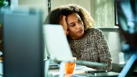 A woman looks bored at her desk in an office. She is glancing off to the side and resting on her hand, not looking at her computer.