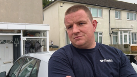 A man with short brown hair and a blue tshirt with his arms folded in front of a white car and some white-painted houses