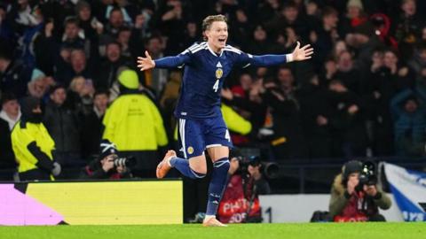 Scotland's Scott McTominay smiling and running with his arms outstretched with fans celebrating in the background.