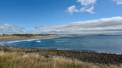 Blue sky and light cloud over the sea with a sandy beach and grassland nearby 