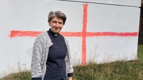 Reverend Rachel Heskin (woman with short hair, blue top and grey cardigan. There is a red cross painted on a white wall behind her). There is also long grass.
