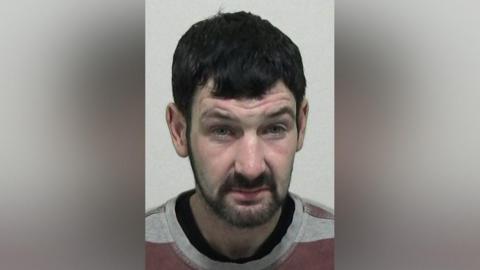 Mugshot of Layton Wood. He has short thick black hair and a black beard, with a dazed expression on his face.