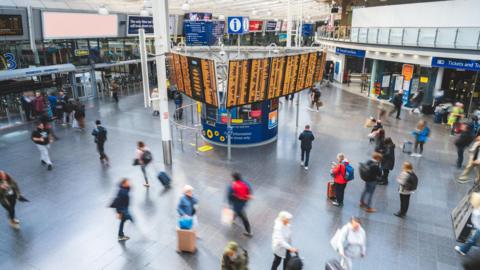 An overhead view of Manchester Piccadilly station and a central departures board lit up with train destinations and times. Around thirty people are crossing the concourse, blurred in the photo due to movement.
