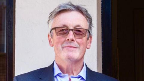 Shane John Gerard Connolly pictured with a neutral expression on his face. He is wearing rectangular shaped glasses, he has grey hair and is wearing a white collar shirt underneath a grey jacket.