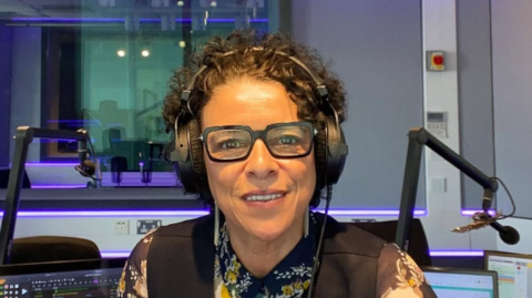 A woman with dark brown hair, and glasses sits in a studio with two microphones behind her. There are two monitors behind her. She is wearing a blue, white and yellow hsirt and a blue waistcoat.