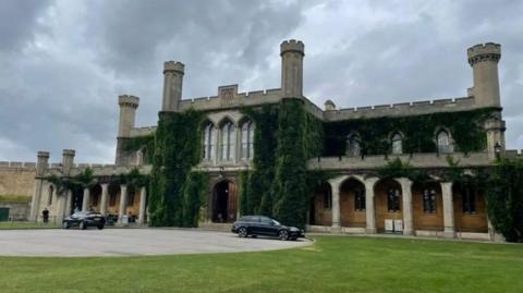 Exterior of Lincoln Crown Court. It is a two-storey Victorian Gothic building with crenellated towers and arches and foliage growing up the walls. In front is a grass lawn and a roadway with two cars parked on it.