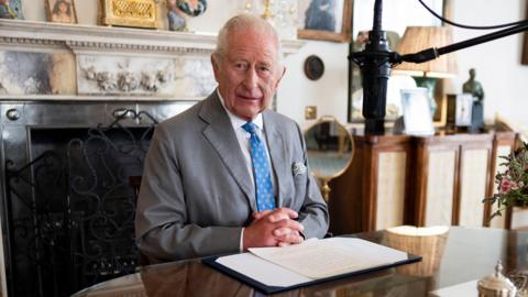 King Charles III wears a grey suit with a striking blue tie with his hands clasped at the foot of a piece of paper containing the words of his VJ Day message in the Morning Room of Clarence House, London. He is sitting in front of a marble-style fireplace. A microphone hovers near to this face.
