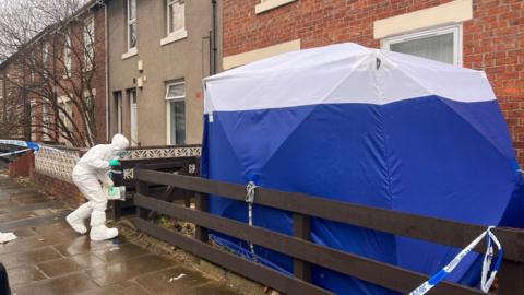 A blue forensic tent outside a red-brick terraced house on a street. Someone in a white haz-mat suit can be seen entering the house.