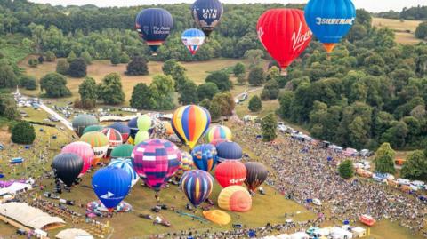 A picture of many hot air ballons on a large field. Some are in the air and others are on the ground. There are large crowds of poeple
