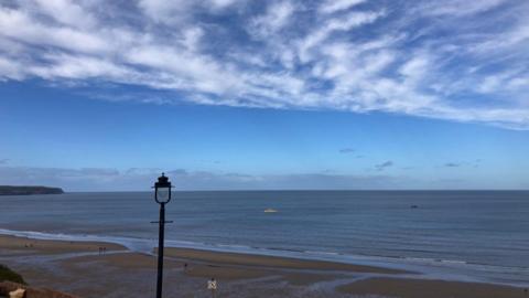 Blue skies streaked with thin white clouds over the sea with the beach and a lamppost in the forefront