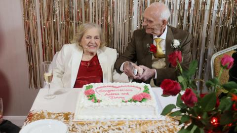 Andrea and Jimmy sat at a table, preparing to cut their wedding cake. Jimmy is smiling at his new bride and she is smiling at the camera.
