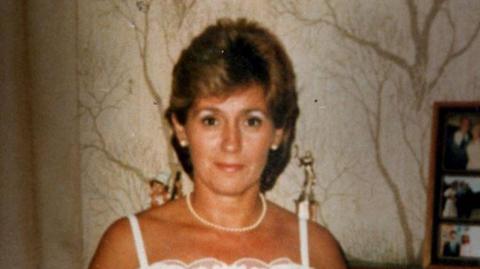 A photograph of Ann Heron. She is wearing a white dress with shoulder straps and a pearl necklace and earrings. She has short brown hair. She is standing inside a house with wallpaper depicting leafless trees. To the right are family photographs.