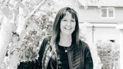 A black and white image of a smiling woman with shoulder-length hair wearing a leather jacket and necklace