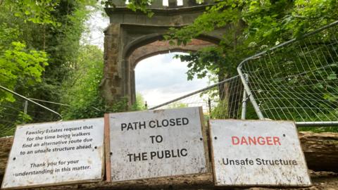 Stone arch with crack in middle stone, surrounded by metal fencing with notices telling walkers to keep out