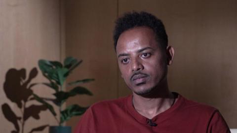 A man with black moustache and hair wearing a maroon t-shirt. He is sitting in a room with wooden cladding walls and a plant in the background.