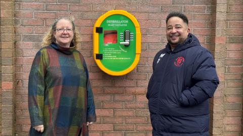 A woman with a multicoloured gown and curly blonde hair stands beside a man with short dark hair wearing a waterproof sports coat. Behind them is a green and yellow plastic case with a red defibrillator inside.