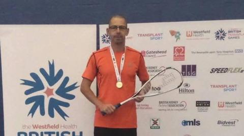 A man in an orange T shirt stands against a media board holding a squash racket. He has a gold medal around his neck. He is wearing rectangular glasses.