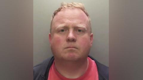 Robert Bush with short, light brown hair is shown in a straight‑on mugshot photograph. He has a neutral expression and is wearing a dark jacket over a red shirt. The background is plain and grey.