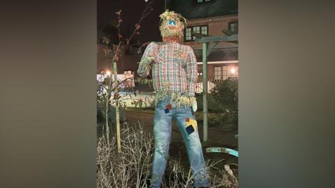 Tex the scarecrow is wearing a tartan shirt and blue jeans with patches on them. He is standing in the middle of a garden arrangement which is in front of buildings.