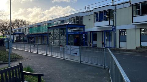 Exterior of QEH hospital front. Modern building painted in blue and yellow. Railings are opposite with a road dissecting the two. Some scaffolding is on the building. 